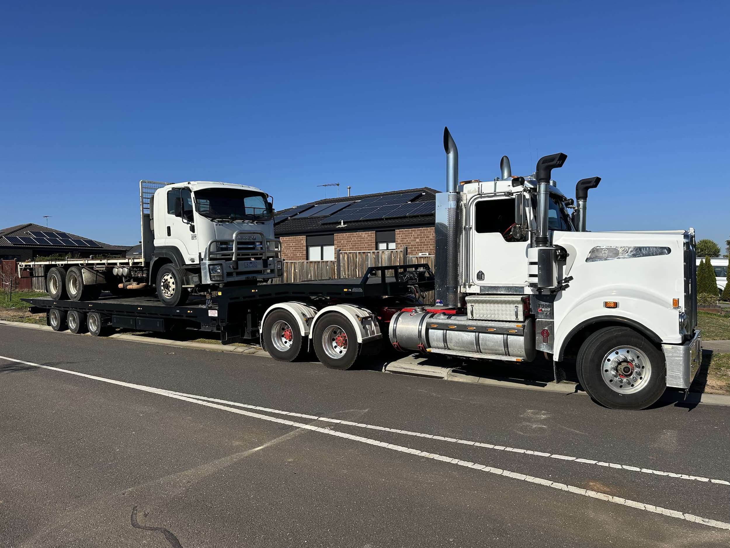 White Kenworth tilt tray truck towing a flatbed truck on suburban street in Melbourne